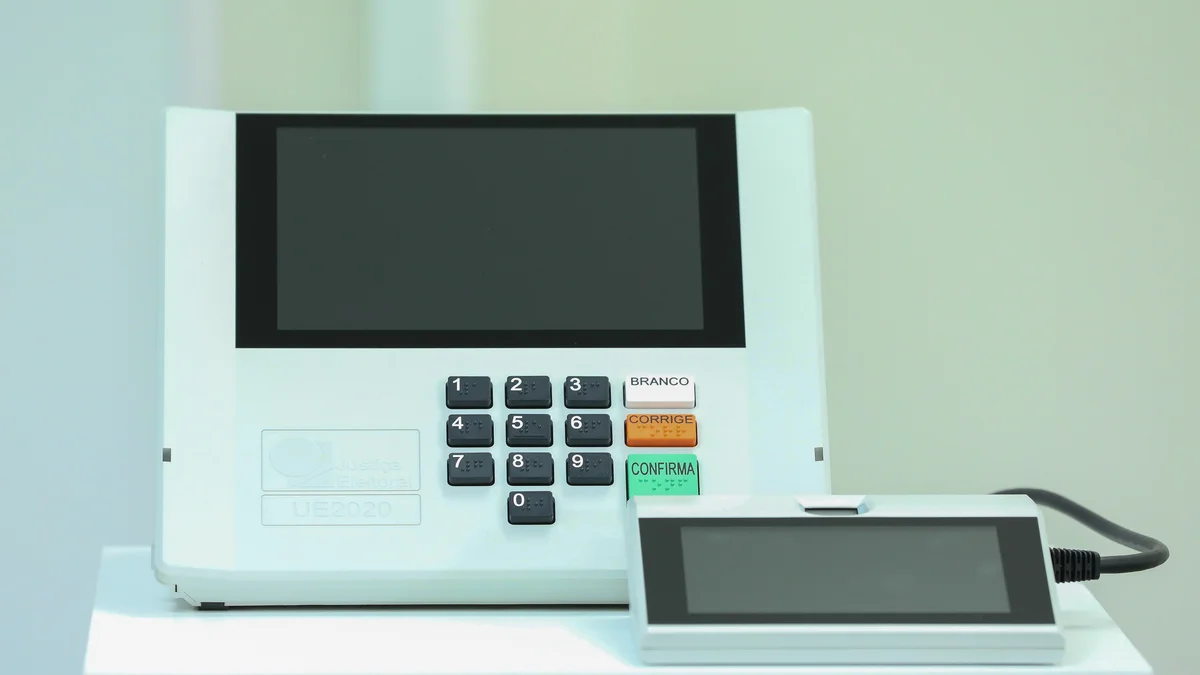 Urna eletrônica brasileira modelo UE2020 com teclado numérico e teclas branco, corrige e confirma, ao lado do terminal do mesário
